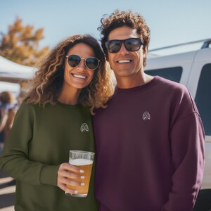 May include: Two people wearing matching sweatshirts with a small embroidered dog design. The woman is wearing a green sweatshirt and holding a glass of beer. The man is wearing a burgundy sweatshirt.
