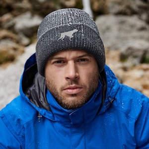 May include: A dark gray knit beanie with a white dog silhouette embroidered on the front. The hat has a folded brim and a textured appearance. The person in the image is wearing a blue jacket.