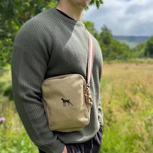 Può includere: Borsa a tracolla in tela beige con silhouette di cane nero. La borsa ha una tracolla a righe con dettagli rosa e bianchi e un dettaglio in corda. La persona indossa un maglione verde e pantaloni neri, in un campo.