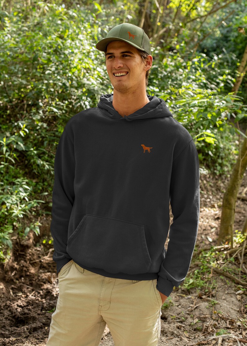May include: A black hooded sweatshirt with a small embroidered brown dog on the left chest. The model is wearing a green baseball cap with a brown dog embroidered on the front.