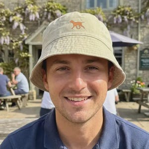 May include: A beige bucket hat with a brown dog silhouette embroidered on the front. The hat is worn by a person wearing a navy blue polo shirt. The background shows a stone building and outdoor seating.