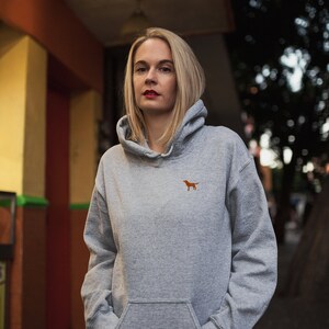 May include: A woman wearing a gray hooded sweatshirt with a small orange dog logo on the front. She is standing in front of a building with a yellow wall and a streetlight.