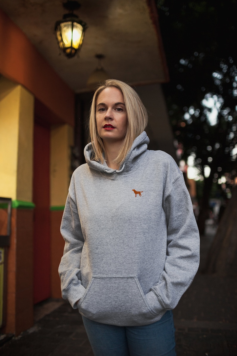 May include: A woman wearing a gray hooded sweatshirt with a small orange dog logo on the front. She is standing in front of a building with a yellow wall and a streetlight.