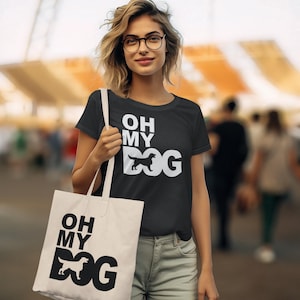 Può includere: T-shirt e borsa tote nere con la scritta bianca "OH MY DOG". La parola "DOG" presenta una silhouette di cane. La donna indossa occhiali e jeans chiari.
