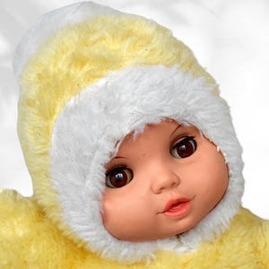 Puede incluir: Una muñeca vintage con un conjunto con capucha amarillo y blanco. La muñeca tiene ojos marrones y un atuendo suave y afelpado. La capucha está adornada con piel sintética blanca. La cara de la muñeca tiene una expresión suave.