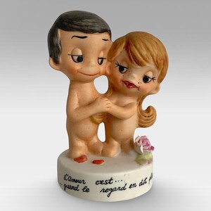May include: A ceramic figurine of a couple embracing, with the man on the left and the woman on the right. The figurine is painted in a realistic style, with the couple's skin tones and facial features visible. The figurine is standing on a white base with the inscription "L'amour c'est... quand le regard en dit plus".