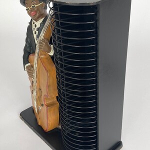 Pu&ograve; includere: Torre porta CD nera con una statuetta di un uomo che suona il contrabbasso. La torre ha una base in legno nero e un mandrino in metallo nero.