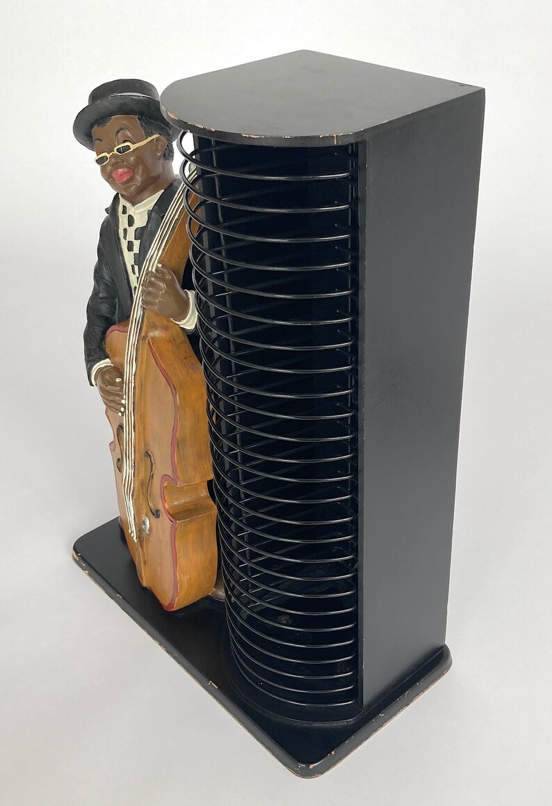 Pu&ograve; includere: Torre porta CD nera con una statuetta di un uomo che suona il contrabbasso. La torre ha una base in legno nero e un mandrino in metallo nero.