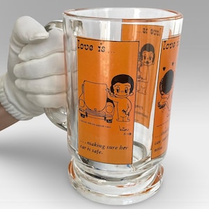 May include: A clear glass beer stein with an orange and black cartoon illustration of a couple. The illustration says "love is... making sure her car is safe." and "kim 5-18".