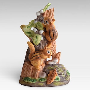 May include: A ceramic figurine of two squirrels on a tree stump. The squirrels have orange fur with black stripes. The tree stump has a detailed bark texture, with green leaves, mushrooms, and an acorn. The base is painted with green and brown tones.