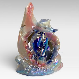 Könnte beinhalten: Eine Keramikfigur mit drei Delfinen in einer Welle, mit irisierendem blauem, rosa und weißem Überzug. Die Delfine sind in einer verspielten Pose, einer springt aus dem Wasser.