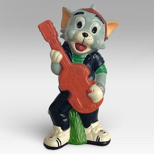 Op de afbeelding: Een grijs en wit Tom en Jerry cartoonfiguur dat een rode elektrische gitaar bespeelt. Het personage draagt een zwart leren jack, een groen shirt en een rode bandana.