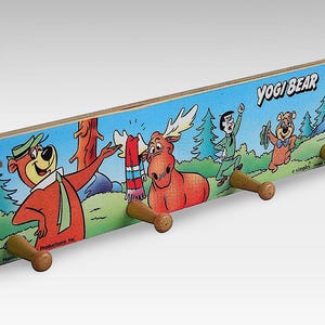 May include: A wooden coat rack featuring Yogi Bear, Boo Boo Bear, and Huckleberry Hound from the cartoon series "Yogi Bear". The rack has four hooks and is decorated with a cartoon scene of the characters in a forest setting.