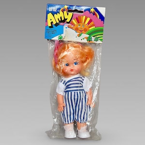 Peut inclure: Une poupée en plastique aux cheveux blonds, aux yeux bleus et vêtue d'une robe à rayures bleues et blanches. La poupée est emballée dans un sac en plastique avec un dos en carton coloré qui dit "Little Amy".