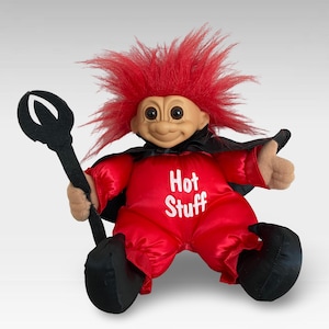 Op de afbeelding: Rode trollpop die een rood duivelskostuum draagt met een zwarte cape en een zwarte drietand vasthoudt. Het kostuum heeft de tekst "Hot Stuff" op de voorkant.