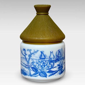 May include: A white ceramic honey pot with a blue and white floral design and a brown lid. The lid is shaped like a thatched roof and has the word "MELI" written on a sign.