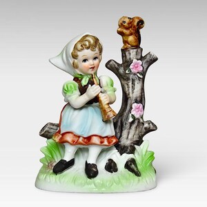 May include: A porcelain figurine of a girl in a white and blue dress playing a flute while sitting on a tree stump with a squirrel perched on top. The figurine is surrounded by green leaves and pink flowers.
