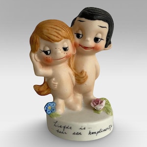 May include: A porcelain figurine of a couple, a girl with blonde hair and a boy with black hair, standing on a base with the text "Liefde is... haar een compliment!"  The figurine is painted in a cartoon style with bright colors.