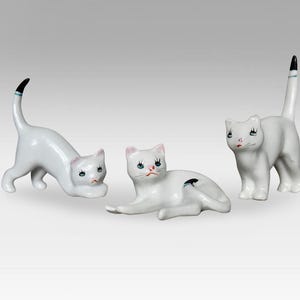 Peut inclure: Trois figurines de chat en porcelaine blanche avec des queues noires et des yeux bleus. Les chats sont dans des poses différentes, l'un est debout, l'autre est couché et l'autre est accroupi.