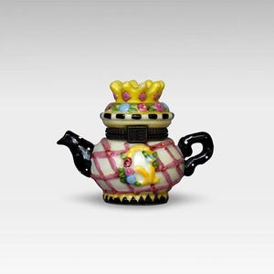 May include: A decorative ceramic teapot with a yellow crown lid. The teapot features a white base with a pink plaid pattern, black spout and handle, and floral accents. The teapot is a unique collectible.
