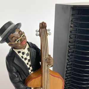 Pu&ograve; includere: Una statuetta di un uomo con un cappello nero e occhiali, che suona un basso marrone e nero. La statuetta &egrave; in piedi accanto a una torre di stoccaggio CD nera.