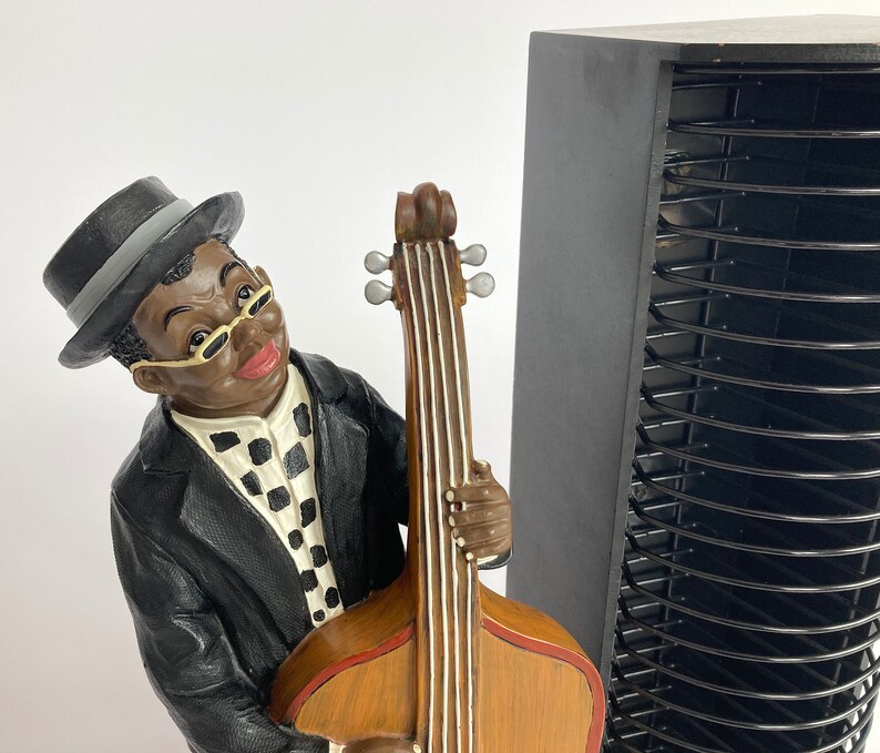 Pu&ograve; includere: Una statuetta di un uomo con un cappello nero e occhiali, che suona un basso marrone e nero. La statuetta &egrave; in piedi accanto a una torre di stoccaggio CD nera.