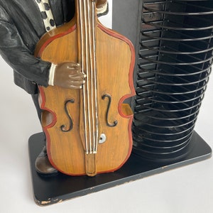 Pu&ograve; includere: Una statuetta di un uomo che suona un contrabbasso, di colore nero e marrone. La statuetta &egrave; in piedi su una base nera con un rack per CD.