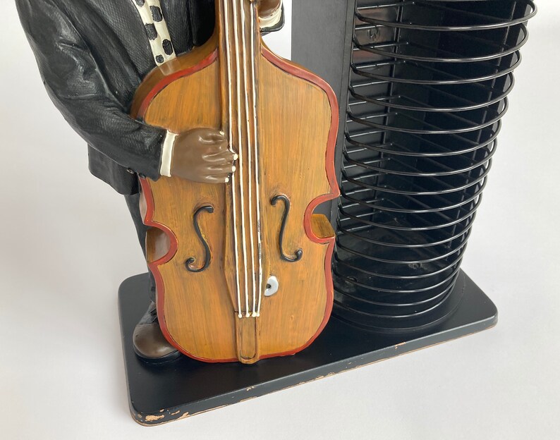 Pu&ograve; includere: Una statuetta di un uomo che suona un contrabbasso, di colore nero e marrone. La statuetta &egrave; in piedi su una base nera con un rack per CD.