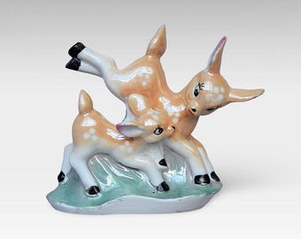 May include: A porcelain figurine of two deer, one standing on its hind legs and the other lying down. The deer are painted in shades of brown and white with black eyes and spots. The figurine is set on a green and white base.