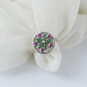 May include: A silver ring featuring a circular head with a floral design. The centre is composed of six marquise-cut green stones arranged in a petal pattern, surrounded by six small round-cut pink stones set within a circular silver frame.