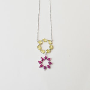 May include: A silver necklace featuring a circular pendant composed of oval-shaped pale yellow gemstones, with a second smaller circular pendant hanging below it, which is composed of teardrop-shaped vibrant magenta gemstones set in silver.