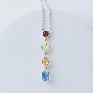 Puede incluir: Un collar de plata con un colgante vertical que presenta cuatro piedras preciosas facetadas en fila. Las piedras incluyen un granate rojo oscuro redondo, un citrino amarillo claro cuadrado, un citrino naranja en forma de pera y un topacio azul claro rectangular.
