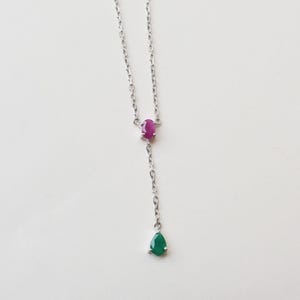 May include: A silver-toned chain necklace featuring a small oval-shaped pink gemstone set in the center, with a delicate vertical drop chain extending downward to a single pear-shaped green gemstone pendant at the very bottom of the piece.