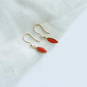 May include: A pair of gold-colored drop earrings with red oval-shaped stones and small clear accent stones. The earrings are displayed on a white, textured fabric background.