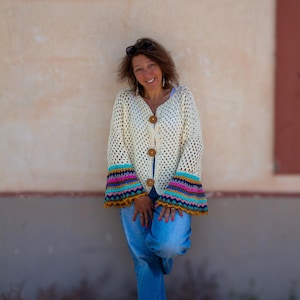 Puede incluir: Cárdigan de crochet color crema con botones grandes de madera y mangas acampanadas coloridas. Las mangas presentan bandas turquesas, rosas, amarillas y naranjas. El cárdigan se combina con vaqueros azules.