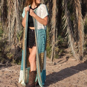 Puede incluir: Un cárdigan largo de ganchillo con diseño patchwork. El cárdigan presenta paneles en crema, turquesa y marrón. La modelo lleva un top negro, pantalones cortos negros y botas de vaquero marrones. El fondo incluye palmeras y suelo arenoso.