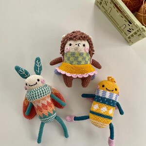May include: Three crocheted stuffed animals: a yellow and blue bee, a brown and yellow hedgehog, and a green and orange moth. The animals are all smiling and have large eyes.