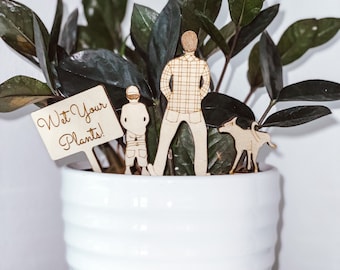 Puede incluir: Una maceta de cerámica blanca con una planta verde y figuras de madera. Las figuras incluyen una persona, un niño y un perro. Un letrero de madera dice "¡Riega tus plantas!"