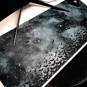 May include: A black and gray desk mat with a galaxy-like design featuring white stars and a flock of bats flying across the surface.