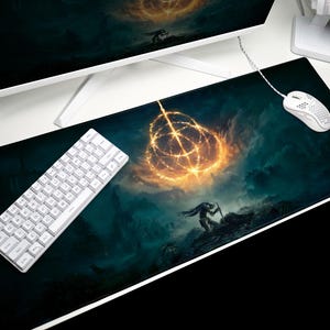 K&ouml;nnte beinhalten: Schwarzes rechteckiges Gaming-Mauspad mit einer Fantasy-Landschaft mit einem leuchtenden goldenen Ring und einer Silhouette. Eine wei&szlig;e Tastatur und Maus befinden sich auf der rechten Seite. Ein Computermonitor ist im Hintergrund.