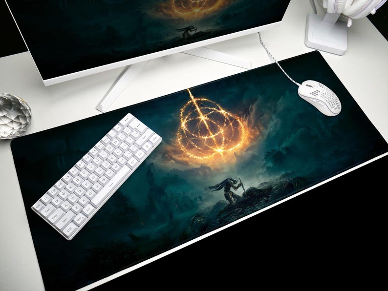 K&ouml;nnte beinhalten: Schwarzes rechteckiges Gaming-Mauspad mit einer Fantasy-Landschaft mit einem leuchtenden goldenen Ring und einer Silhouette. Eine wei&szlig;e Tastatur und Maus befinden sich auf der rechten Seite. Ein Computermonitor ist im Hintergrund.