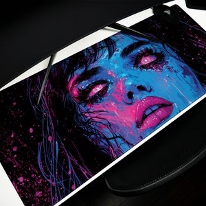May include: A black and blue desk mat with a colorful abstract portrait of a woman with pink lips and glowing eyes. The background is black with pink and blue splatters.