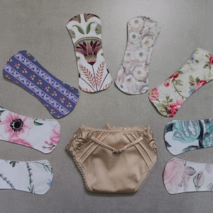 Puede incluir: Un surtido de compresas de tela reutilizables y un par de bragas de satén beige. Las compresas presentan varios diseños florales y estampados en colores como rosa, azul y verde. La ropa interior tiene un lazo decorativo.