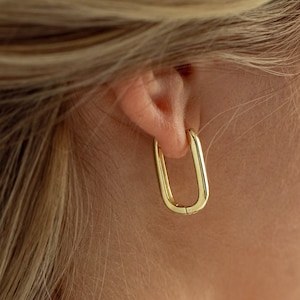 May include: Gold hoop earrings with a unique U-shaped design. The earrings are shown in a close-up view on a person's ear.