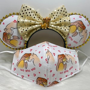 May include: A themed accessory set featuring a Simba design. Includes mouse ears with a gold sequin bow and a face mask. The mask and ears are white with a Simba illustration and the text "we are one."