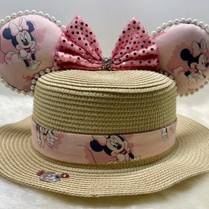Puede incluir: Un sombrero de paja con una cinta de temática Minnie Mouse rosa y blanca y un lazo rosa con lentejuelas. El sombrero tiene orejas de Minnie Mouse con tela rosa y blanca y ribete de perlas.