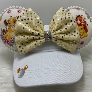 May include: A white visor with a gold glitter bow and two fabric mouse ears. The ears feature a floral design with images of Simba and Pumbaa from The Lion King.