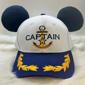 May include: White baseball cap with blue brim and black Mickey Mouse ears. The cap has a gold anchor and the word "CAPTAIN" embroidered on the front. The brim has gold laurel wreaths.