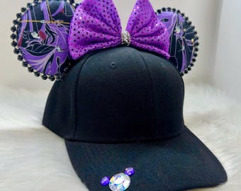 Op de afbeelding: Een zwarte baseballpet met een paarse glitter strik en paarse en zwarte gepatroneerde stoffen muisoren. De oren hebben zwarte kralen rond de randen. De pet heeft een klein strass ontwerp aan de voorkant.