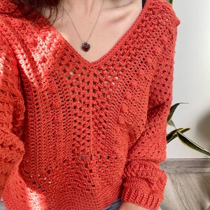 Puede incluir: Un suéter de crochet naranja con cuello en V profundo y mangas largas. El suéter tiene un tejido abierto y texturizado. Se ve un collar plateado con un colgante en forma de corazón. La persona lleva vaqueros azules.